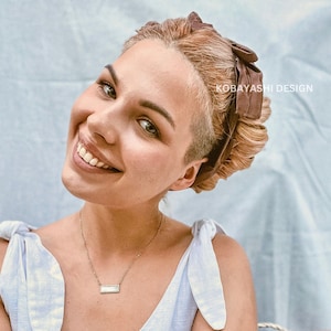 May include: A woman with short brown hair styled with a brown bow. She is wearing a light blue sleeveless top with a white and blue pattern. She is smiling and wearing a silver necklace with a rectangular pendant.