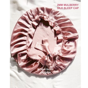 Op de afbeelding: Een roze slaapmuts van 2 mm zijde van de moerbeiboom. De muts heeft een gerimpeld ontwerp en een strik aan de achterkant. De tekst "Kobayashi Design" is op de muts gedrukt.