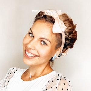 May include: A woman with blonde hair wearing a white shirt with black polka dots and a pink velvet headband with a bow.