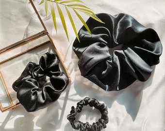 May include: Three black satin scrunchies of different sizes. The largest scrunchie is lying flat on a white surface. The two smaller scrunchies are in a glass box with a gold frame.