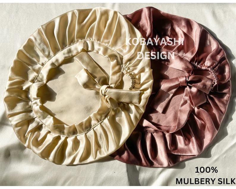 Op de afbeelding: Twee satijnen slaapmutsen, een in lichtbeige en de andere in stoffig roze. Beide mutsen hebben een geplooide vorm en een strik aan de bovenkant. De tekst "KOBAYASHI DESIGN" is gedrukt op de roze muts. De tekst "100% MULBERRY SILK" is gedrukt onder de mutsen.