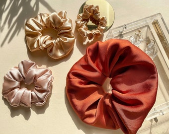 May include: Three satin scrunchies in different colors: peach, beige, and burnt orange. The scrunchies are arranged on a light beige surface.