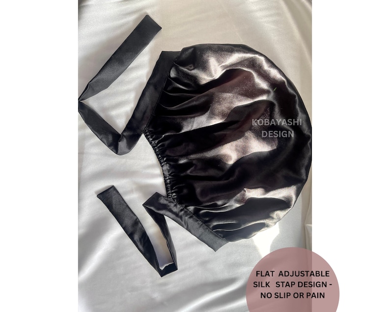 Op de afbeelding: Zwarte satijnen slaapmuts met verstelbare bandjes. De muts is ontworpen om haarbreuk en pluis te voorkomen. "Kobayashi Design" is op de muts gedrukt. "Flat Adjustable Silk Strap Design - No Slip or Pain" is onder de muts gedrukt.