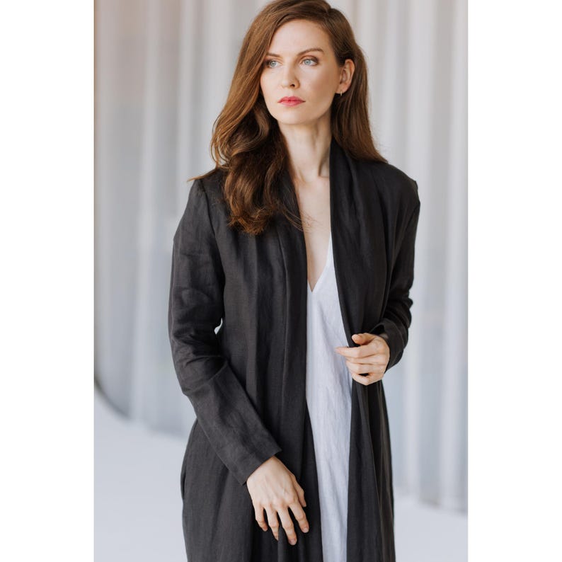K&ouml;nnte beinhalten: Dunkel anthrazitfarbene lange Leinenstrickjacke mit drapiertem Kragen. Die Strickjacke wird &uuml;ber einem wei&szlig;en Kleid getragen. Die &Auml;rmel sind lang und der Stoff scheint leicht zu sein. Der Gesamtstil ist minimalistisch und elegant.
