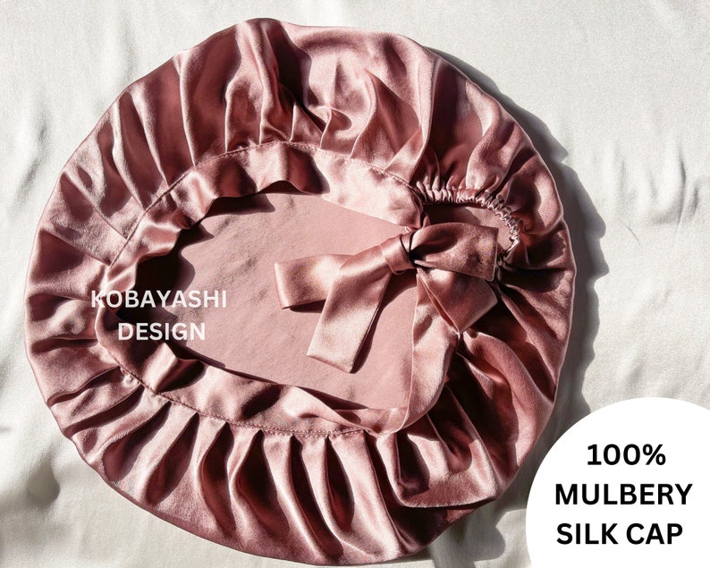 Op de afbeelding: Een stofrijke roze zijden slaapmuts met een strik. De muts is gemaakt van 100% moerbeizijde en is ontworpen om het haar te beschermen tijdens het slapen.