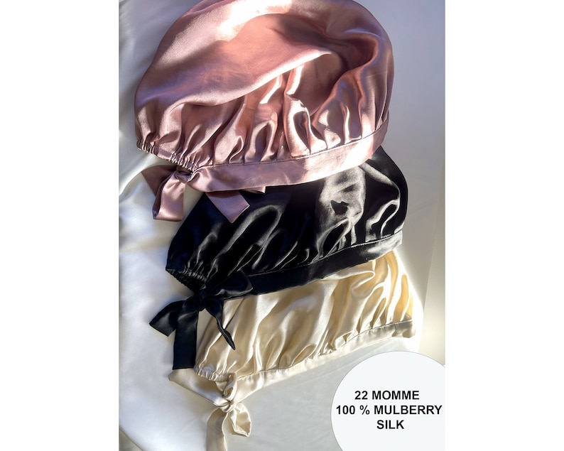 Op de afbeelding: Drie slaapmutsen van zijde in de kleuren roze, zwart en cr&egrave;me. De mutsen zijn gemaakt van 100% moerbeizijde en zijn op elkaar gestapeld. De tekst "22 Momme 100% Mulberry Silk" is zichtbaar onderaan de afbeelding.