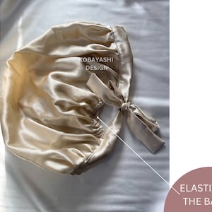 Op de afbeelding: Een beige satijnen slaapmuts met een elastiek aan de achterkant. De muts heeft een strik aan de achterkant en de woorden "KOBAYASHI DESIGN" staan ​​gedrukt op de voorkant.