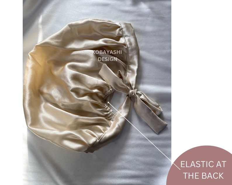 Op de afbeelding: Een beige satijnen slaapmuts met een elastiek aan de achterkant. De muts heeft een strik aan de achterkant en de woorden "KOBAYASHI DESIGN" staan ​​gedrukt op de voorkant.