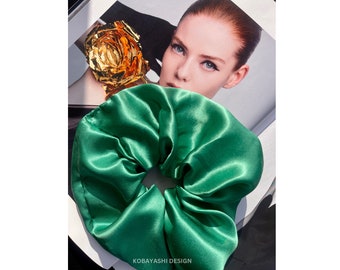 May include: A green satin scrunchie hair tie. The scrunchie is made of soft, silky fabric and has a gathered, ruffled look.
