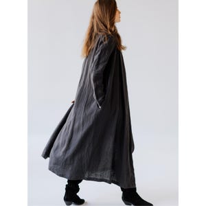 K&ouml;nnte beinhalten: Ein langer, anthrazitfarbener Leinenmantel mit entspannter Passform. Der Mantel hat lange &Auml;rmel, Taschen und eine flie&szlig;ende Silhouette. Das Model tr&auml;gt schwarze Stiefel. Dieses Kleidungsst&uuml;ck eignet sich zum &Uuml;berziehen und bietet eine minimalistische &Auml;sthetik.