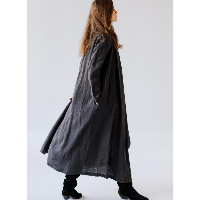K&ouml;nnte beinhalten: Ein langer, anthrazitfarbener Leinenmantel mit entspannter Passform. Der Mantel hat lange &Auml;rmel, Taschen und eine flie&szlig;ende Silhouette. Das Model tr&auml;gt schwarze Stiefel. Dieses Kleidungsst&uuml;ck eignet sich zum &Uuml;berziehen und bietet eine minimalistische &Auml;sthetik.