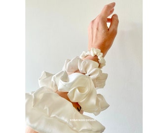 May include: Three white satin scrunchies, two are stacked on top of each other and one is worn on a wrist. The scrunchies are made of soft, silky fabric and have a gathered, ruffled look. The text "KOBAYASHI DESIGN" is visible at the bottom of the image.