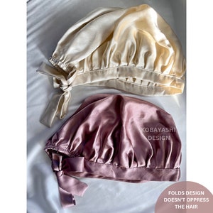 Op de afbeelding: Twee satijnen slaapmutsen, een in lichtbeige en de andere in stoffig roze. Beide mutsen hebben een gerimpeld ontwerp en een strik aan de achterkant. De tekst "Kobayashi Design" is gedrukt op de roze muts. De tekst "Folds Design Doesn't Oppress The Hair" is gedrukt onder de mutsen.