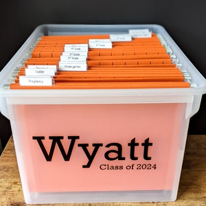 May include: A clear plastic storage box with orange hanging folders labeled with different grade levels. The box has a pink label on the front that says "Wyatt Class of 2024".
