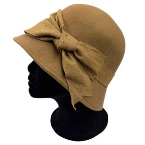 May include: A tan felt cloche hat featuring a large, decorative bow detail on the side. The hat has a rounded crown and a short, downward-sloping brim. The fabric bow is made of the same tan material and is positioned prominently on the side.