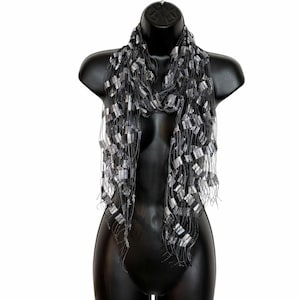 May include: A black and silver scarf featuring an open-weave, ladder-like construction with metallic silver rectangular accents interspersed throughout the dark, thin, thread-like material, creating a textured, layered, and lightweight appearance.