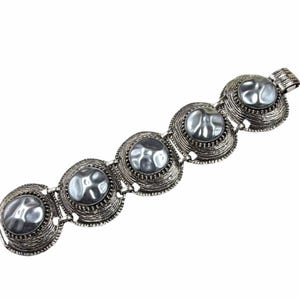 May include: A silver-toned link bracelet featuring five circular segments. Each segment contains a smooth, grey, domed center piece surrounded by a textured, ridged metal border. The bracelet is connected by small metal hinges and includes a clasp.