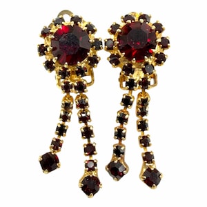 May include: A pair of gold-tone clip-on earrings featuring a large, round, deep red faceted stone at the center, surrounded by a halo of smaller red stones. Two dangling chains of square-cut red stones extend from the base of each earring.