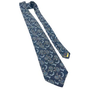 Puede incluir: Una corbata de seda azul con un patrón floral blanco y azul claro. La corbata tiene una punta puntiaguda y un ancho estrecho. El nombre de la marca "ROBERT ALBOIS" es visible en la parte posterior de la corbata.