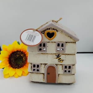 May include: A cream ceramic house-shaped candle holder with a heart-shaped window and a bee decoration. The house has multiple windows and a brown door. A yellow sunflower is next to the house.