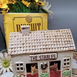 Può includere: Una casa decorativa in ceramica con la scritta "THE STABLES" sopra le porte della stalla. La casa ha un tetto strutturato e presenta teste di cavallo nelle aperture della stalla. La casa è color crema con porte della stalla rosse e piante in vaso verdi.