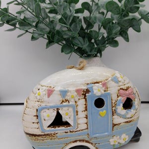 Può includere: Un vaso in ceramica a forma di camper, bianco e blu. Il camper ha una porta, finestre ed elementi decorativi come fiori e un fiocco rosa. Rami verdi e frondosi emergono dalla parte superiore del vaso.