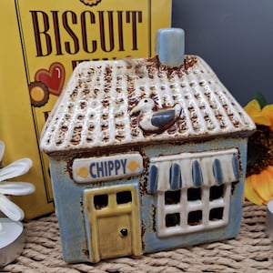 May include: A ceramic house-shaped candle holder with a blue facade, a brown roof, and a yellow door. The word "CHIPPY" is displayed above the door. A decorative duck is on the roof. The background includes a yellow book with the word "BISCUIT".