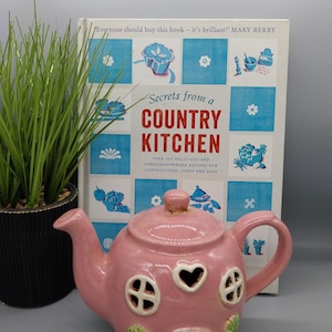 Könnte beinhalten: Eine rosa Keramik-Teekanne in Form eines Hauses mit herzförmigen und kreisförmigen Aussparungen. Die Teekanne steht neben einem Kochbuch mit dem Titel "Secrets from a Country Kitchen" und einer Topfpflanze.
