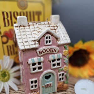 May include: A pink ceramic house-shaped candle holder with a white roof and the word "BOOKS" above the door. The door is light blue with a heart-shaped cutout and the word "OPEN". Decorative flowers and a cat are on the side.