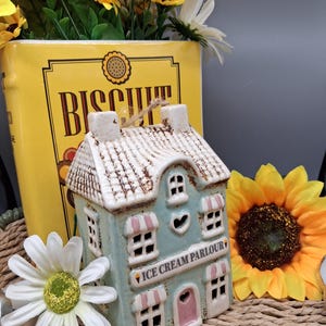 Może przedstawiać: Ceramiczna figurka lodziarni z jasnoniebieską fasadą, różowymi akcentami i napisem "ICE CREAM PARLOUR". Figurka ma biały dach i jest otoczona słonecznikami i stokrotkami.