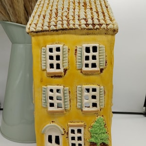 May include: A yellow ceramic house with white trim and a green plant in a brown pot. The house has windows with white frames and green shutters. The roof is brown with white tiles.