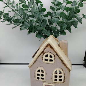 Może przedstawiać: Ceramiczny świecznik w kształcie domu z jasnofioletowym szkliwem i różowymi drzwiami. Dom ma po dwa okna z każdej strony i komin na szczycie.
