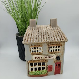 Op de afbeelding: Een keramische kandelaar in de vorm van een huis met een grijze buitenkant, bruin dak en de tekst "POST OFFICE" op een bord. Het huis heeft een rode deur en ramen. Een groene plant in een zwarte pot staat op de achtergrond.