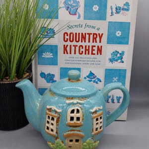 Può includere: Una teiera in ceramica azzurra a forma di casa, con finestre e una porta. La teiera è davanti a un libro di cucina intitolato "Secrets from a Country Kitchen". Una pianta verde è a sinistra.