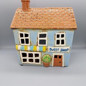 May include: A blue and brown ceramic house shaped like a small shop with a sign that reads "Sweet Shop". The house has a brown roof, a chimney, two windows, a door, and a small plant in a pot.