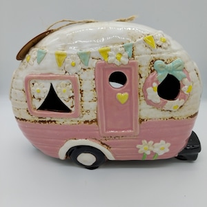 May include: A pink ceramic camper figurine with white accents, decorated with flowers and bunting. The camper has a pink door with a heart cutout and a wreath on the back.
