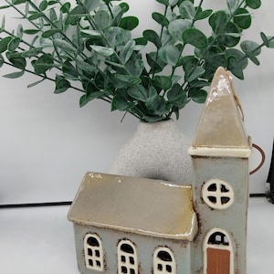 May include: A ceramic church figurine with a gray and brown glaze. The church has a tall steeple and three windows. The figurine is sitting on a white surface.
