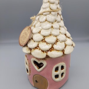 Pode incluir: Uma casa de cerâmica rosa com telhado branco e detalhes marrons. A casa tem uma janela em forma de coração e uma pequena porta. O telhado é texturizado com pequenas formas redondas. A casa é decorada com um laço de corda marrom.