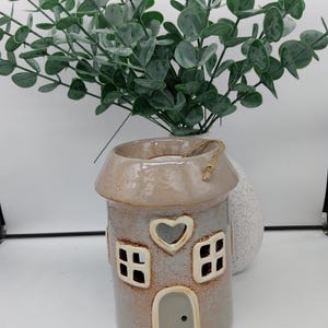 May include: A ceramic house shaped candle holder with a heart cutout and two windows. The house is painted in a light brown color with a white trim. The candle holder is sitting on a white surface.