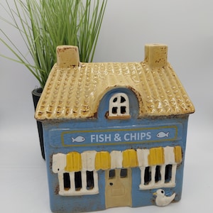 Könnte beinhalten: Keramikfigur in Form eines Hauses mit blauer Fassade und gelbem Dach. Das Schild trägt die Aufschrift "FISH & CHIPS" mit Fischillustrationen. Mit kleiner Tür, Fenstern und einer dekorativen Möwe. Eine Topfpflanze befindet sich im Hintergrund.