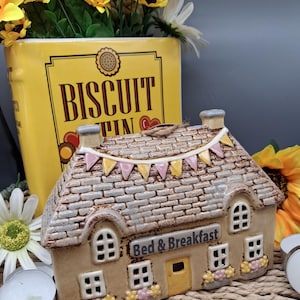 May include: A ceramic Bed & Breakfast cottage candle holder with a brick-effect roof and a string of pink and yellow bunting. A yellow biscuit tin and sunflowers are in the background. The cottage has a yellow door.