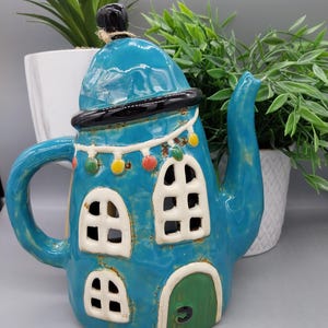 Può includere: Una teiera in ceramica blu turchese a forma di casa con tetto nero, finestre bianche e una porta verde. La teiera ha una ghirlanda di luci colorate intorno al tetto e un piccolo pomo nero in cima.