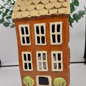 Può includere: Una statuetta di casa in ceramica con corpo marrone, finestre bianche e tetto beige. La casa ha due piccoli alberi in vaso su entrambi i lati della porta d'ingresso.