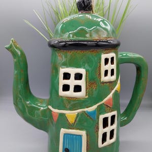 May include: A green ceramic teapot shaped like a house with a blue door, white windows, and colourful bunting. The teapot has a black rim and a black knob on the lid.