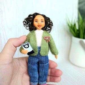 May include: A small doll with curly dark brown hair, wearing a white shirt, a green knit cardigan with a pink heart-shaped pin, blue denim jeans, and pink shoes. The doll holds a miniature black and white smartphone with a panda face design.