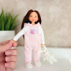 May include: A handmade doll wearing a pink polka-dot onesie with a white hooded top and fuzzy slippers. The doll has brown hair and is holding a small white stuffed bunny.