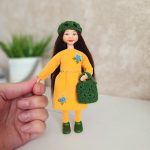 May include: A handmade doll with long brown hair, wearing a mustard yellow dress with blue floral accents, a green crocheted hat, and green shoes. The doll is holding a green crocheted handbag. The doll is a miniature art piece.