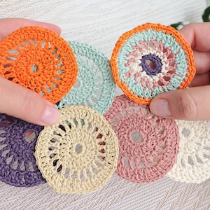 Può includere: Una collezione di sottobicchieri colorati all'uncinetto in varie tonalità di arancione, verde, viola, beige, rosa e bianco. I sottobicchieri hanno una forma circolare con un design traforato. Un sottobicchiere è tenuto in una mano.
