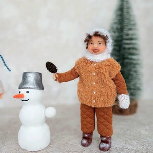 May include: A handmade doll wearing a brown fuzzy coat and matching pants, a white hat, and mittens. The doll is holding a small pine cone and standing next to a snowman with a silver bucket hat.