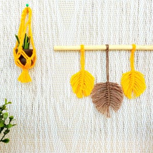 Può includere: Un appendiabiti per piante in macramè giallo con una piccola pianta in vaso e tre decorazioni a forma di foglia in macramè giallo e marrone. Gli oggetti sono appesi a una parete bianca strutturata.